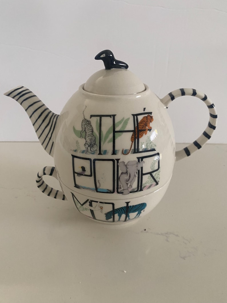 Puede incluir: Tetera de cer&aacute;mica color crema con asa y boquilla a rayas blancas y negras. La tetera presenta ilustraciones de tigres, cebras y un elefante, con las palabras "THE POOR MOM" impresas en la parte delantera. Una figura de p&aacute;jaro negro est&aacute; en la tapa.
