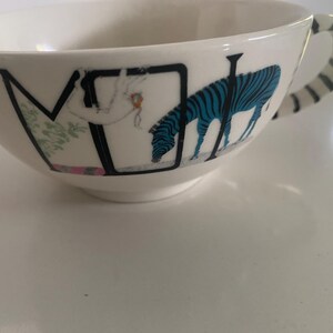 Puede incluir: Taza de t&eacute; de cer&aacute;mica blanca con una ilustraci&oacute;n de cebra negra y azul. La taza presenta un asa con rayas blancas y negras. La taza tiene la letra "M" y una figura de una persona en el lateral.