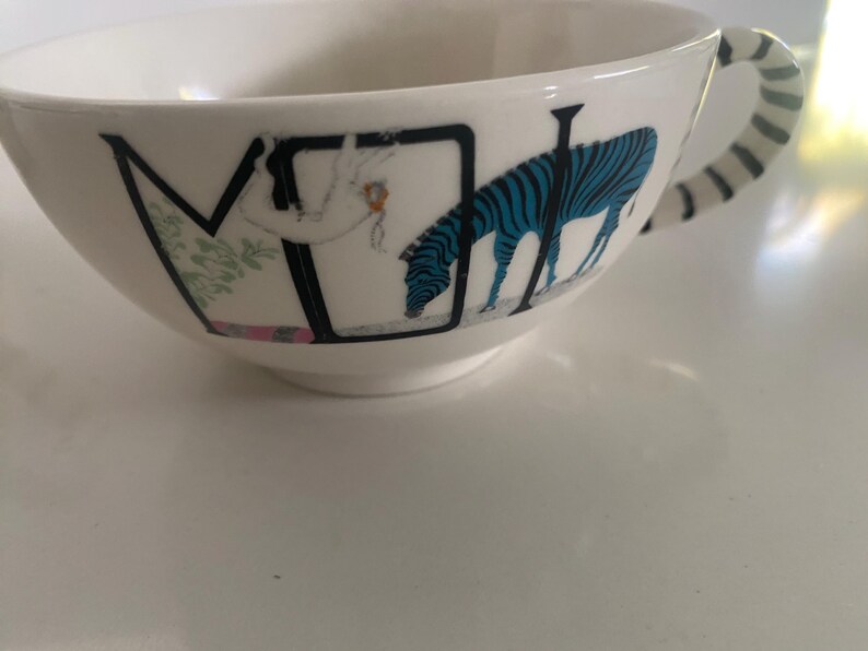 Puede incluir: Taza de t&eacute; de cer&aacute;mica blanca con una ilustraci&oacute;n de cebra negra y azul. La taza presenta un asa con rayas blancas y negras. La taza tiene la letra "M" y una figura de una persona en el lateral.