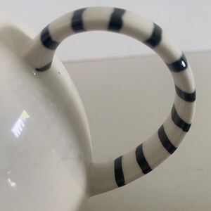 Puede incluir: Taza de cer&aacute;mica blanca con asa curva. El asa est&aacute; decorada con rayas negras y blancas alternas. La taza tiene un acabado brillante y un fondo liso.