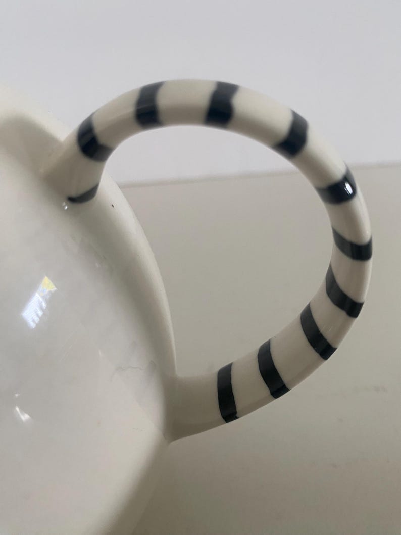 Puede incluir: Taza de cer&aacute;mica blanca con asa curva. El asa est&aacute; decorada con rayas negras y blancas alternas. La taza tiene un acabado brillante y un fondo liso.