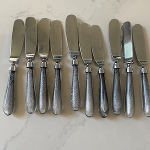 May include: A collection of ten butter knives. Each knife has a stainless steel blade and a teardrop-shaped handle. The handles are clear with a silver, crackled pattern. The knives are displayed in a row.