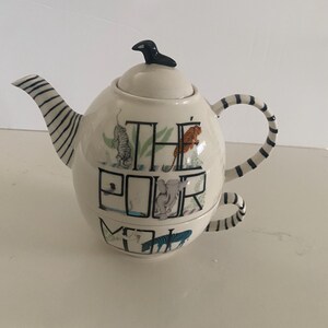 Puede incluir: Tetera de color crema con asa y boquilla a rayas blancas y negras. La tetera tiene un p&aacute;jaro negro en la tapa y las palabras "THE POUR ME" con ilustraciones de animales. La tetera est&aacute; sobre una superficie blanca.