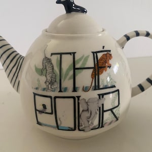 Puede incluir: Tetera de cer&aacute;mica blanca con un asa de tapa en forma de p&aacute;jaro negro. La tetera presenta la palabra "THE ROAR" con ilustraciones de un tigre, un elefante y otros animales. El asa y el pico tienen rayas blancas y negras.