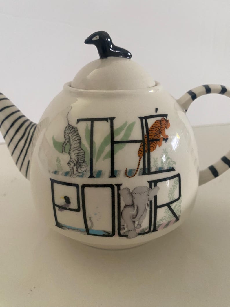 Puede incluir: Tetera de cer&aacute;mica blanca con un asa de tapa en forma de p&aacute;jaro negro. La tetera presenta la palabra "THE ROAR" con ilustraciones de un tigre, un elefante y otros animales. El asa y el pico tienen rayas blancas y negras.