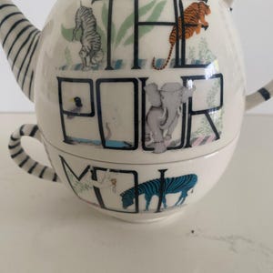 Puede incluir: Tetera de cer&aacute;mica blanca con taza a juego. La tetera presenta las palabras "THE POUR ME" con ilustraciones de animales, incluyendo un tigre y una cebra. El asa y el pico tienen rayas azules y blancas.
