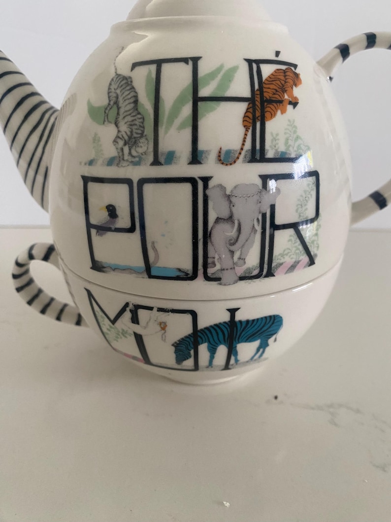 Puede incluir: Tetera de cer&aacute;mica blanca con taza a juego. La tetera presenta las palabras "THE POUR ME" con ilustraciones de animales, incluyendo un tigre y una cebra. El asa y el pico tienen rayas azules y blancas.
