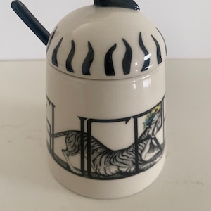 May include: A ceramic sugar bowl with a black bird-shaped lid handle and a matching spoon. The bowl is white with black vertical lines around the top and a black and white illustration of a tiger. The spoon is black.
