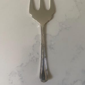 Puede incluir: Un tenedor de servir de metal plateado con tres dientes anchos y planos, y un mango decorativo con detalles de ranuras verticales. El utensilio tiene un acabado pulido y reflectante con una cabeza de forma distintiva que se ensancha.