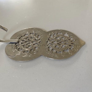 Pode incluir: Um design de metal prateado, decorativo, ornamentado, vazado, em forma de gancho. O design tem a forma de dois corações.