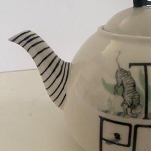 Puede incluir: Tetera de cer&aacute;mica color crema con un asa a rayas blancas y negras y un adorno negro. La tetera presenta un dise&ntilde;o caprichoso con una ilustraci&oacute;n de tigre y la palabra "TEA".