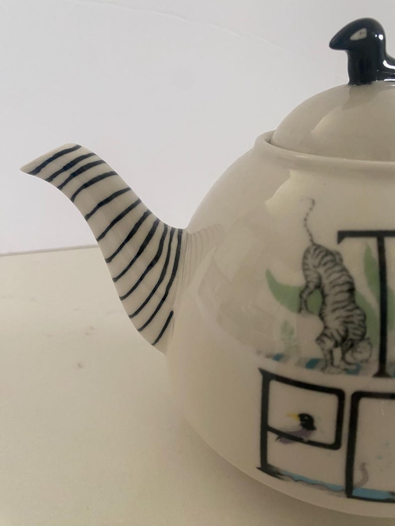 Puede incluir: Tetera de cer&aacute;mica color crema con un asa a rayas blancas y negras y un adorno negro. La tetera presenta un dise&ntilde;o caprichoso con una ilustraci&oacute;n de tigre y la palabra "TEA".