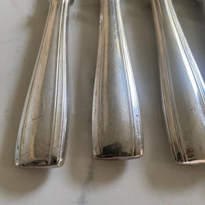 Könnte beinhalten: Nahaufnahme mehrerer silberfarbener Messer. Die Griffe haben ein geriffeltes Design und eine rechteckige Basis. Die Klingen sind oben sichtbar. Die Messer sind in einer Reihe angeordnet, was auf ein Besteckset hindeutet.