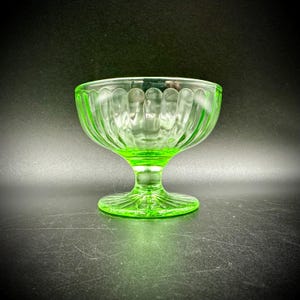May include: A vintage green glass dessert dish with a ribbed design. The bowl sits atop a short stem and a circular base. The glass has a bright, translucent appearance, and the dish is set against a dark background.