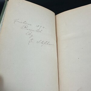 May include: Open book with handwritten inscription: "Christmas 1894. Presented By E. Stephan." The text is in cursive on a cream-colored page. The book's spine is visible, with a green and white patterned design.