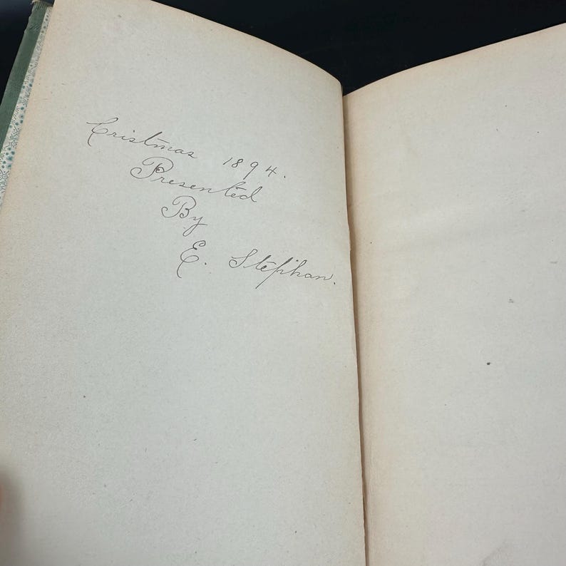 May include: Open book with handwritten inscription: "Christmas 1894. Presented By E. Stephan." The text is in cursive on a cream-colored page. The book's spine is visible, with a green and white patterned design.