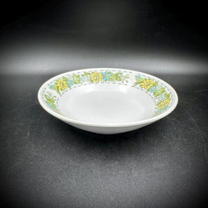 May include: White ceramic bowl with a floral pattern in yellow, blue, and green around the rim. The bowl has a shallow, rounded shape, suitable for serving soups or salads. The bowl is set against a dark background.