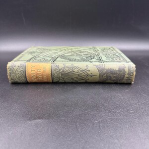 May include: A vintage book titled "Vanity Fair" by Thackeray. The book has a light green cover with ornate designs and a gold label on the spine. The book's edges are slightly worn, suggesting its age.