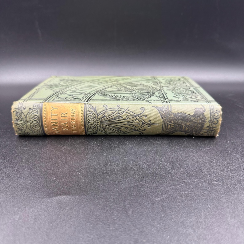 May include: A vintage book titled "Vanity Fair" by Thackeray. The book has a light green cover with ornate designs and a gold label on the spine. The book's edges are slightly worn, suggesting its age.