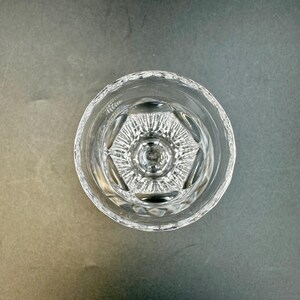 May include: Clear, cut-glass drinking glass with a detailed geometric pattern at the base. The glass has a thick, textured rim and a clear, transparent appearance. The glass is empty and sits on a gray surface.