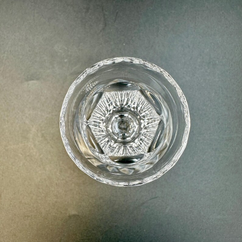May include: Clear, cut-glass drinking glass with a detailed geometric pattern at the base. The glass has a thick, textured rim and a clear, transparent appearance. The glass is empty and sits on a gray surface.