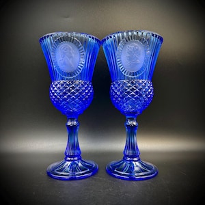 May include: Two cobalt blue goblets. Each goblet features a detailed portrait within a circular frame on the bowl. The bowls have a diamond pattern, and the stems are fluted. The goblets sit on a round base.