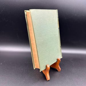 May include: A vintage book with a light green cover and a wooden spine, displayed on a small wooden stand. The book's pages have a warm, aged tone, and the cover shows slight wear. The background is a dark, solid color.