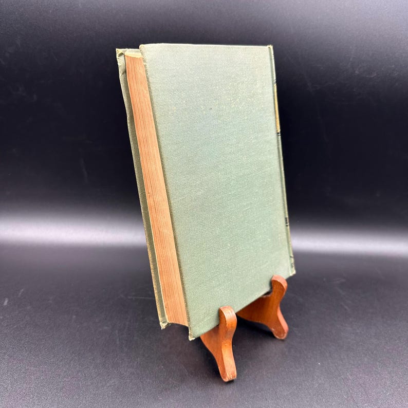 May include: A vintage book with a light green cover and a wooden spine, displayed on a small wooden stand. The book's pages have a warm, aged tone, and the cover shows slight wear. The background is a dark, solid color.
