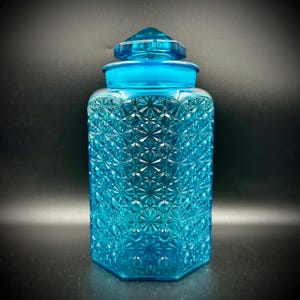 May include: A turquoise glass jar with a geometric pattern and a matching lid. The jar has a hexagonal base and a diamond-like texture, creating a sparkling effect. The lid is a smaller version of the jar's shape.