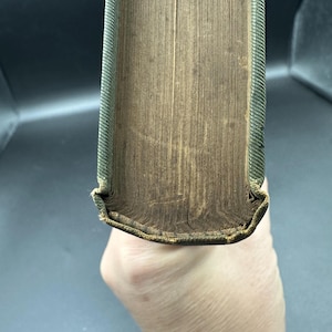 May include: Close-up of an antique book's aged pages and spine. The pages are a warm, faded brown, with visible wear and tear. The spine is a textured green with gold accents, showing signs of age and use.