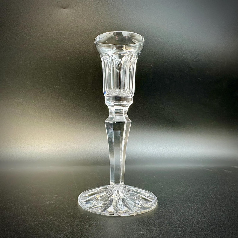 May include: A clear crystal candlestick holder with a round base and a tall, slender stem. The top portion has vertical grooves, and the base features a starburst design. The candlestick is designed to hold a single candle.
