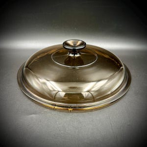 May include: A clear, domed glass lid with a black knob. The lid has a circular design and a silver rim. The glass has a slight amber tint. The lid is designed for cookware.