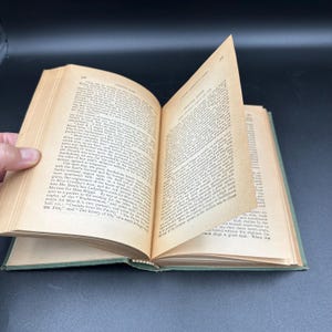 May include: An open vintage book with aged, yellowed pages. The book's cover is a muted green, and the pages are filled with printed text. The book is slightly open, revealing the text and the binding.
