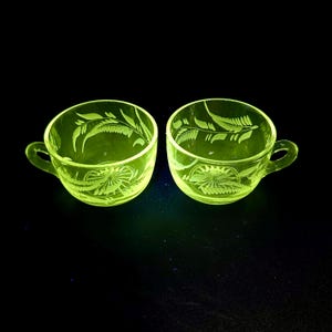 May include: Two vintage, glowing green glass teacups with handles. Each cup features etched fern and floral designs. The cups are set against a black background, highlighting their intricate details and unique color.