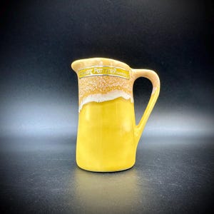 May include: A yellow ceramic pitcher with a handle and spout. The top portion has a textured, off-white and tan design, with the text "Wilart Pottery" in a rectangular label. The pitcher is on a dark background.