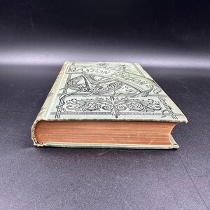 May include: A vintage book with a light green cover and ornate black detailing. The cover features the text "Caxton Edition" and decorative illustrations. The book's pages are a warm, aged orange color, suggesting its age and use.