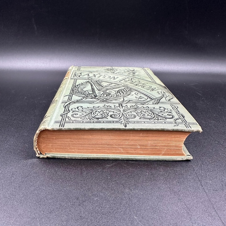 May include: A vintage book with a light green cover and ornate black detailing. The cover features the text "Caxton Edition" and decorative illustrations. The book's pages are a warm, aged orange color, suggesting its age and use.