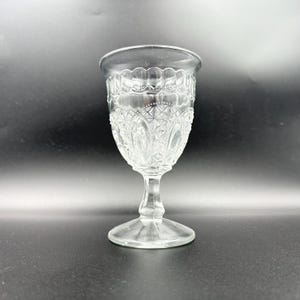May include: A clear, vintage-style glass goblet with a decorative pattern. The goblet features a detailed design on the bowl and a stem. The glass is transparent, allowing light to pass through, and is suitable for serving beverages.
