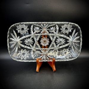 May include: A clear, rectangular glass serving dish with a starburst and floral pattern. The dish is supported by a wooden stand. The design features various star and flower shapes, creating a decorative and elegant appearance.