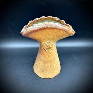 May include: A handmade ceramic mushroom-shaped sculpture with a textured, speckled surface. The sculpture features a wide, scalloped top with a light green rim and a conical base. A circular emblem is embossed on the stem. The color is a warm, earthy tan.