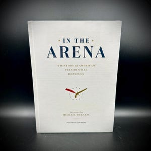 May include: A hardcover book titled "IN THE ARENA" with navy blue text and a clock graphic. The book's subtitle reads "A HISTORY of AMERICAN PRESIDENTIAL HOPEFULS." The cover features the author's name, Michael Dukakis.