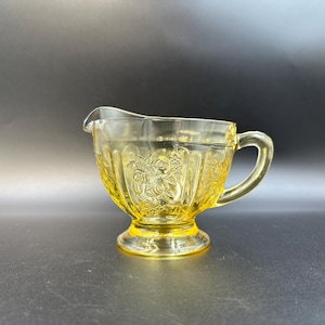 May include: A vintage yellow glass creamer with a floral pattern. The creamer has a handle and a spout for easy pouring. The base is a pedestal design. The glass is a transparent yellow color.