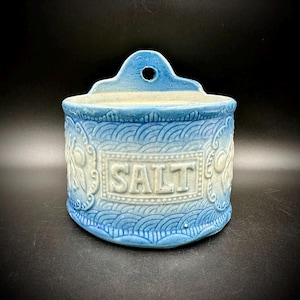 May include: A vintage blue ceramic salt cellar with a decorative wave pattern and the word "SALT" in a rectangular frame. The salt container has a hole at the top for hanging.