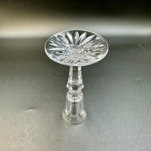 May include: A clear, cut-glass pedestal cake stand with a round top and a faceted stem. The top features a starburst design, and the base is a rounded shape. The stand is made of crystal and is suitable for displaying desserts.
