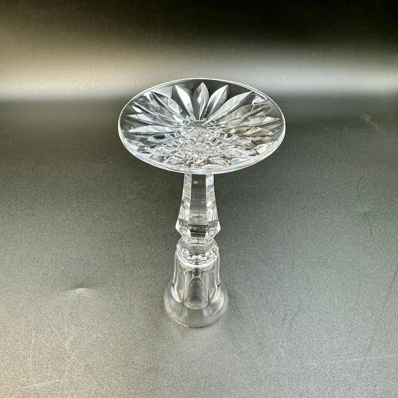 May include: A clear, cut-glass pedestal cake stand with a round top and a faceted stem. The top features a starburst design, and the base is a rounded shape. The stand is made of crystal and is suitable for displaying desserts.