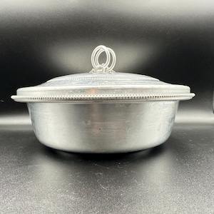 May include: A silver-colored metal serving bowl with a lid. The lid has a looped handle and a beaded edge. The bowl has a rounded shape and a matching beaded edge. The background is black.