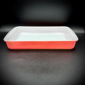May include: A rectangular baking dish with a vibrant coral-orange exterior and a smooth, glossy white interior. The vessel features rounded corners and a wide, flat rim, presenting a clean, functional design for oven-to-table food preparation.