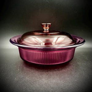 May include: A vintage, ruby-colored glass casserole dish with a matching lid. The dish has a ribbed exterior and a clear, smooth rim. The lid features a small, decorative handle. The dish is designed for baking and serving.
