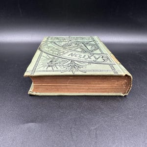 May include: An antique book with a light green cover featuring the text "Baxton's Dictionary" in a decorative font. The book's pages are aged, and the edges are worn, suggesting its age and use.