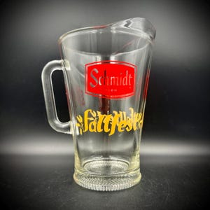May include: Clear glass beer pitcher with a handle. The pitcher has a red and white logo that says "Schmidt Beer" and yellow text that says "Fattfest". The pitcher is empty and sits on a dark surface.